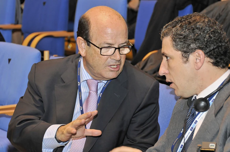 two men in business attire are sitting on a blue chair