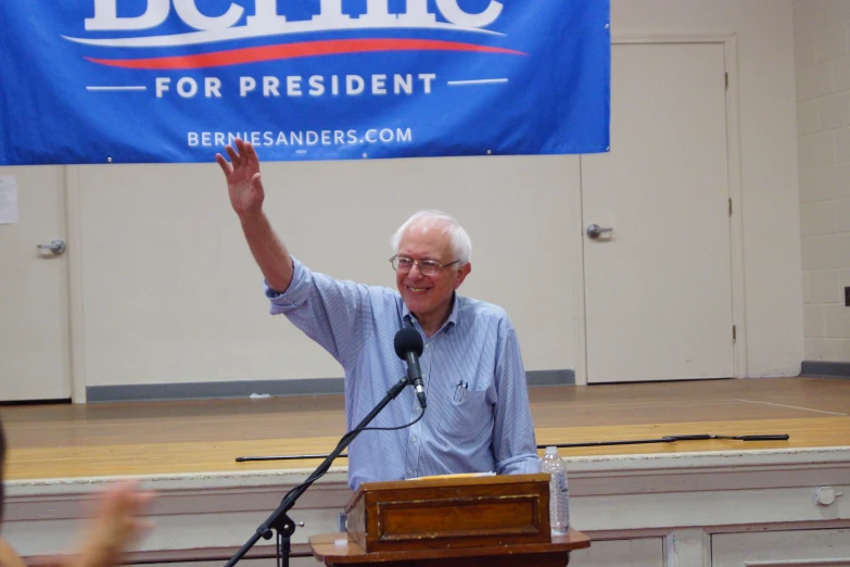 man in glasses standing at podium holding up the president sign