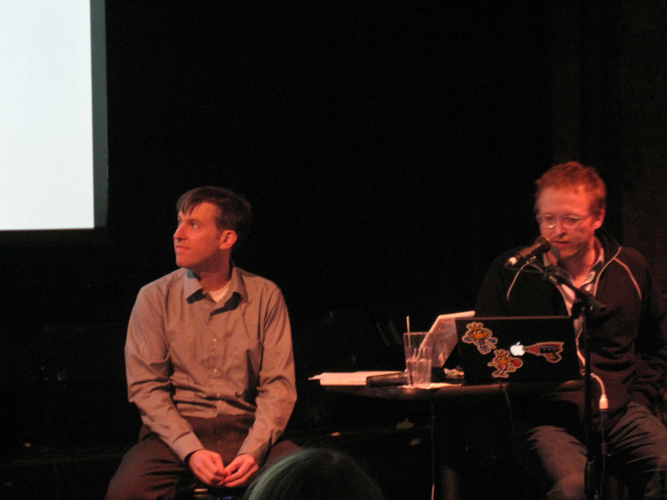 two men sitting on stage one has a microphone and is talking