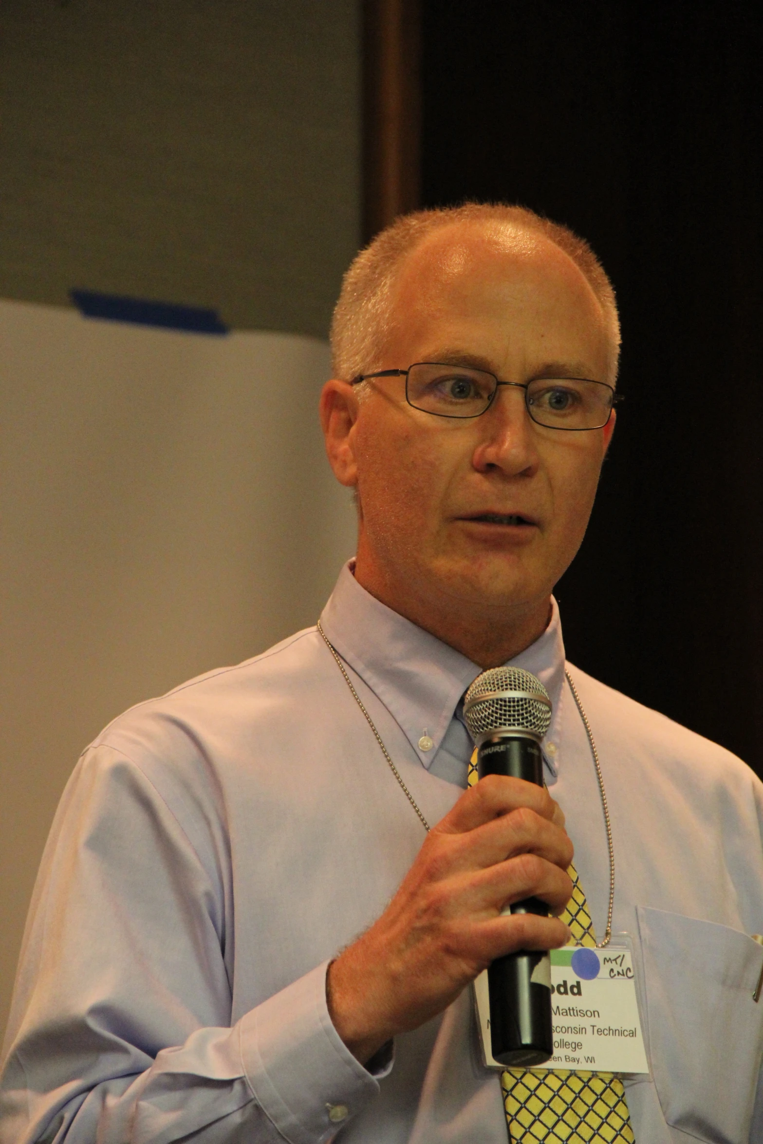 an older man with glasses, holding a microphone and wearing a blue shirt