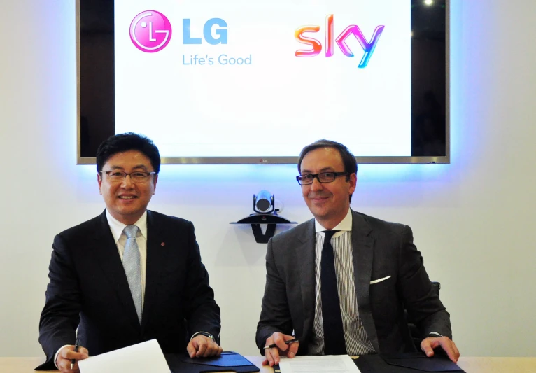 two men sit at a table in front of a projector screen and sign