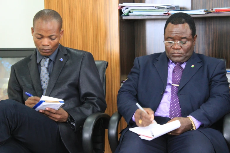 two men in suits sitting down while checking a paper