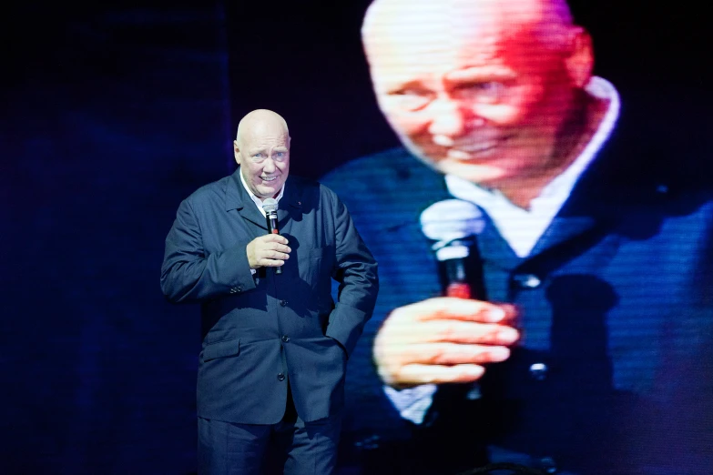 an older man stands next to a microphone in front of a painting of a man