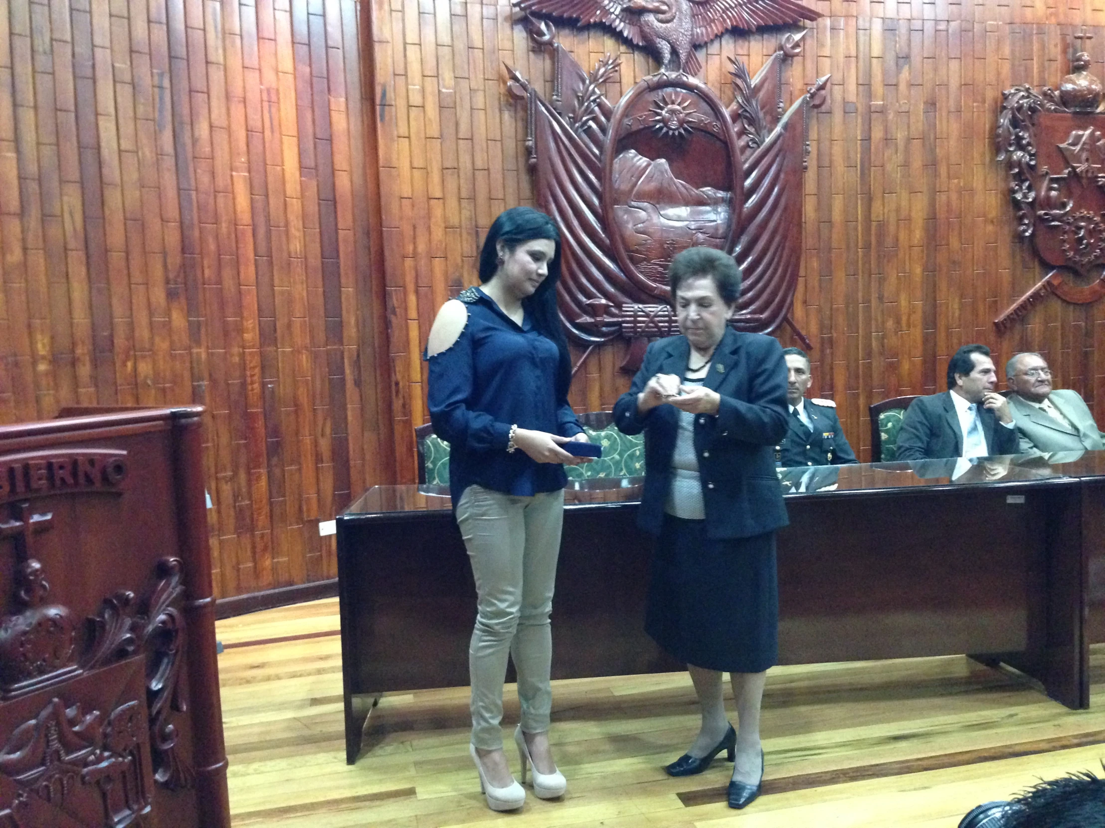 two woman stand in front of an ornate desk while one woman looks at her phone