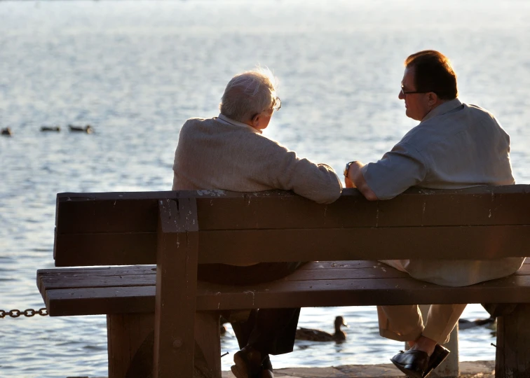 an elderly man and woman sitting on a bench with ducks swimming in the water