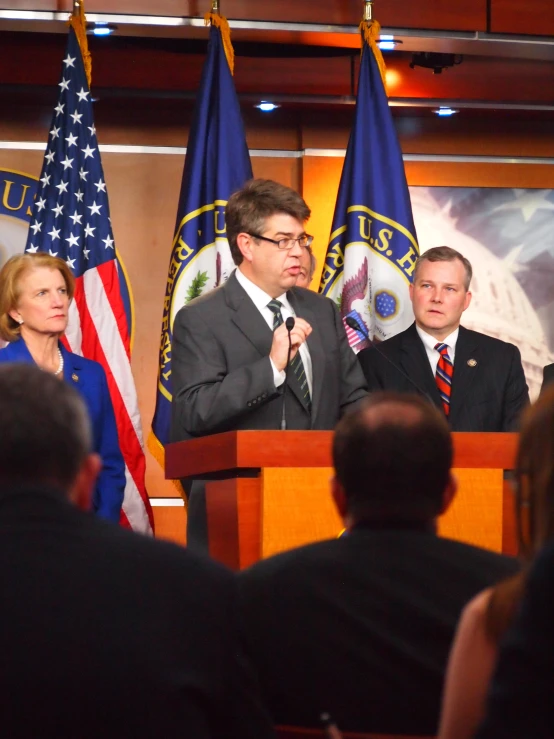 people in business attire stand at microphones behind flags