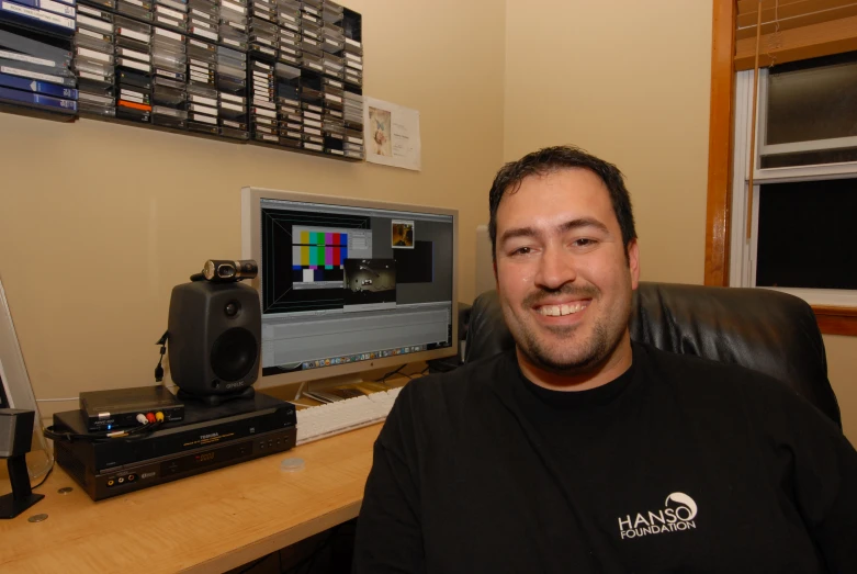 a person is smiling in front of a desk with two computers and a sound recorder on it