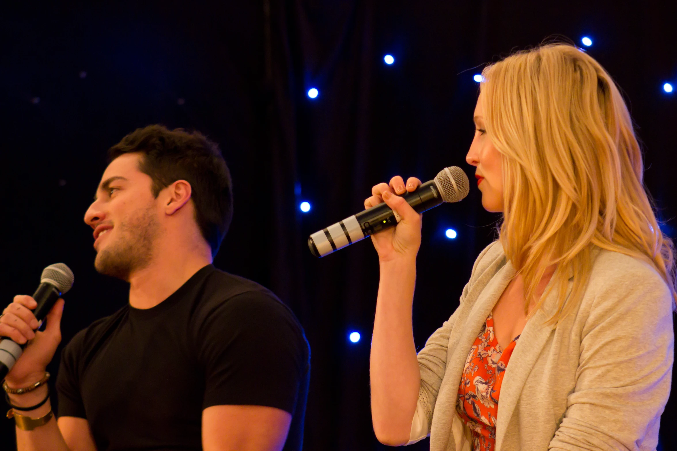 two people stand next to each other on stage and one holds a microphone in front of their head