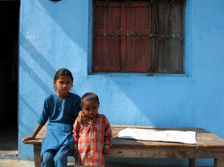 two children sitting on a wooden bench in front of a blue building