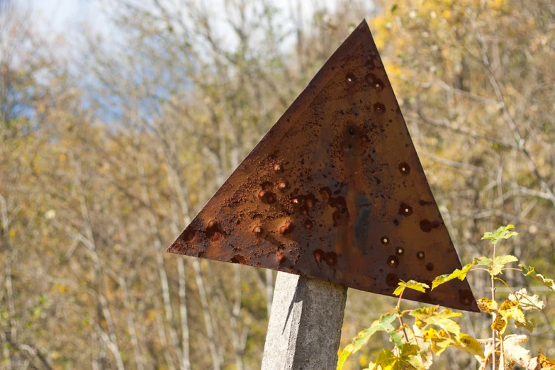 a triangular rusted metal sign sitting next to some trees