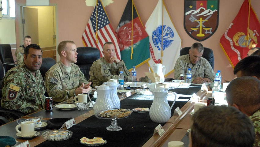 military men sitting at a conference table in front of flags