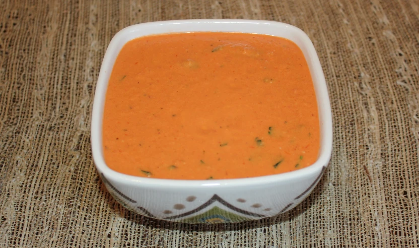 a square bowl of tomato soup on a mat