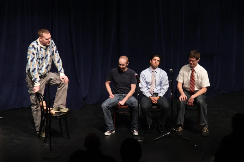 four men sit on stage in front of dark curtains