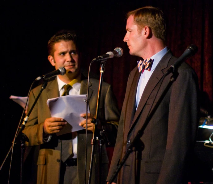 two men standing behind microphones while one sings