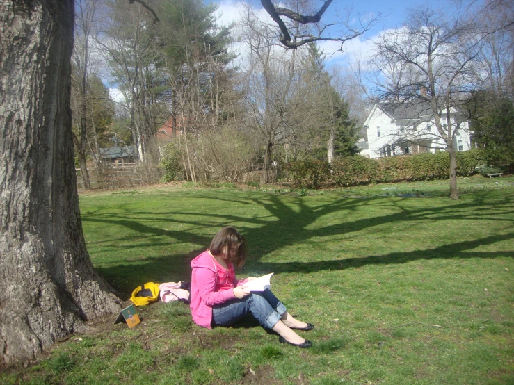 a woman sitting in the grass reading a book next to a tree