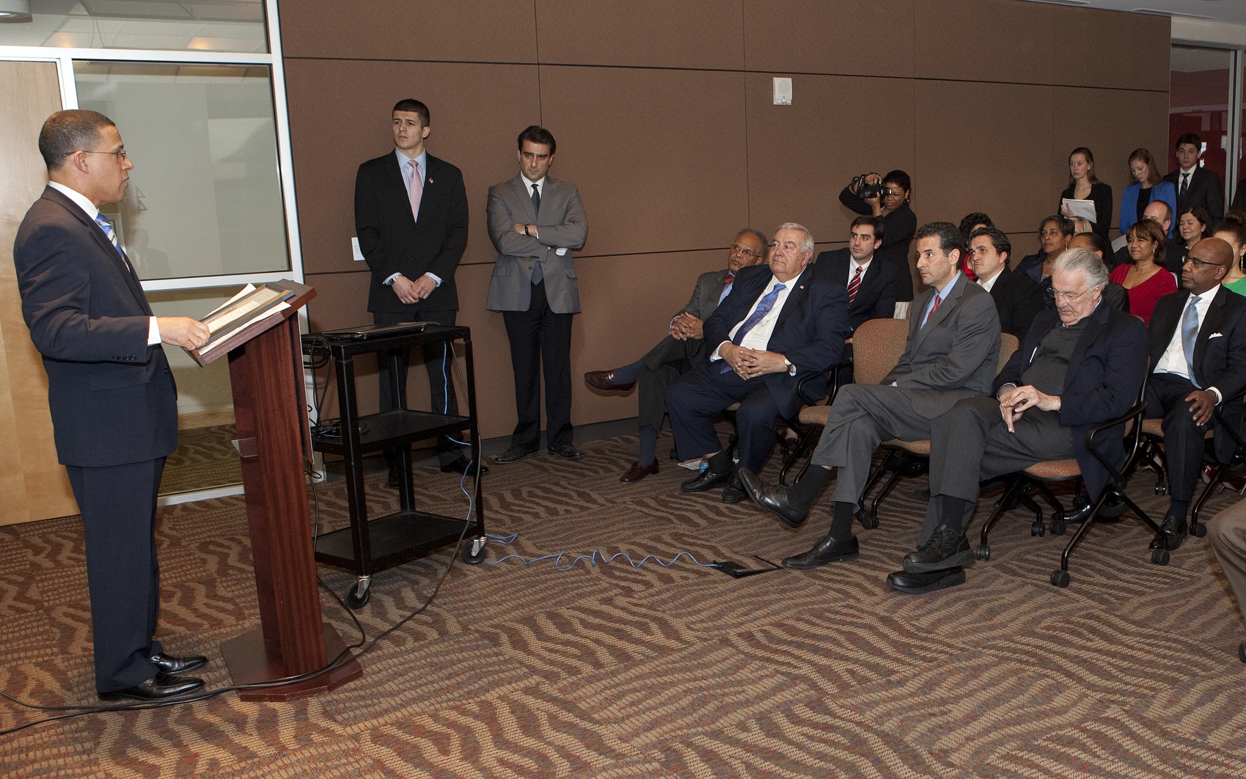 man speaking at podium with many sitting on chairs