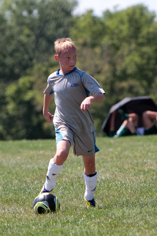 a boy in grey uniform kicking soccer ball on grassy field