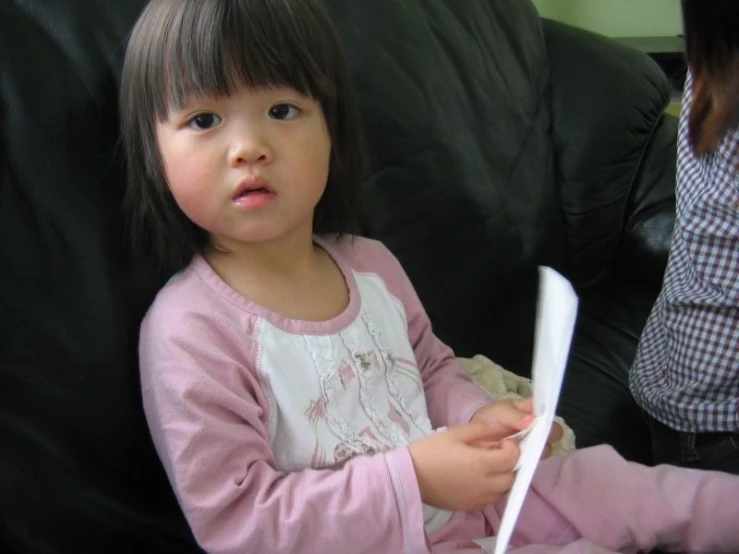 a little girl in a dress sitting on a couch and holding a paper