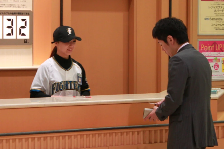 a man in a grey suit and hat stands at the counter talking to a woman in a white shirt and black baseball cap