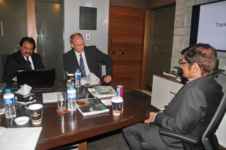 three men sitting around a table in front of a laptop