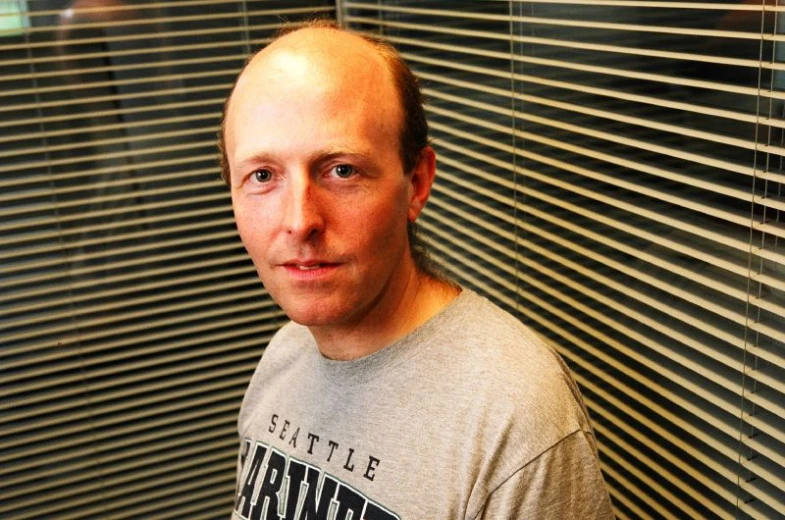 a bald headed man standing near blinds, looks at the camera