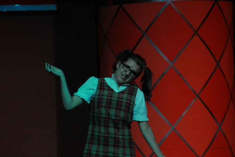 a girl wearing plaid vest and glasses is on stage with one hand up in the air