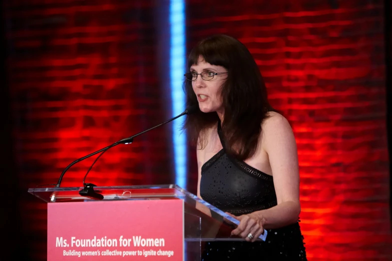 woman standing at a podium wearing glasses and speaking