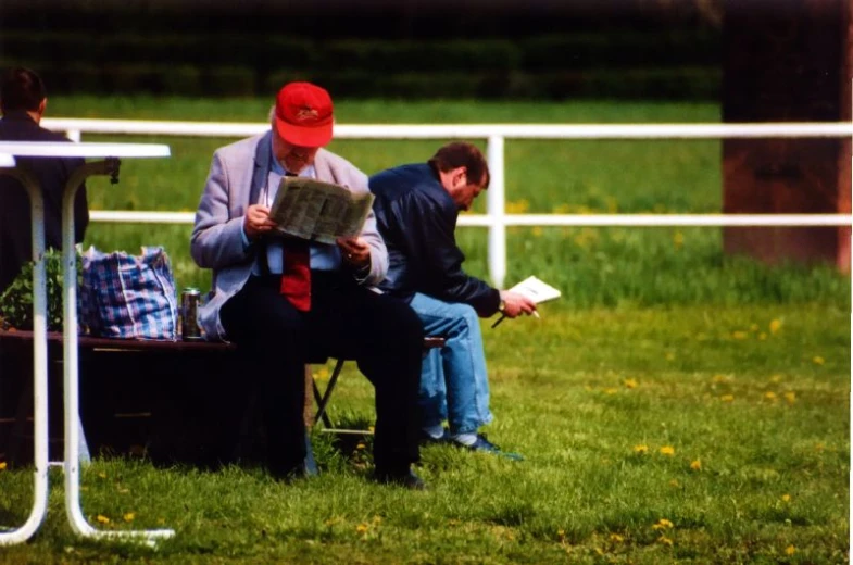 two men are in the grass reading while one holds a paper