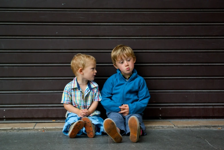 two boys sitting on the ground while one is looking at the camera
