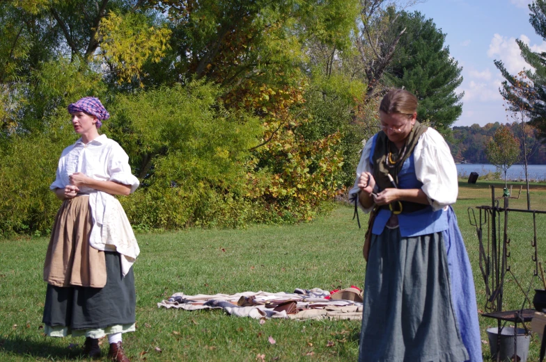 two women in period clothing and hats stand near an open fire pit