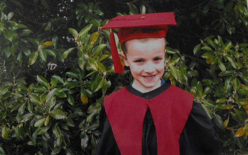 a boy wearing a red hat and gown and standing in front of some bushes