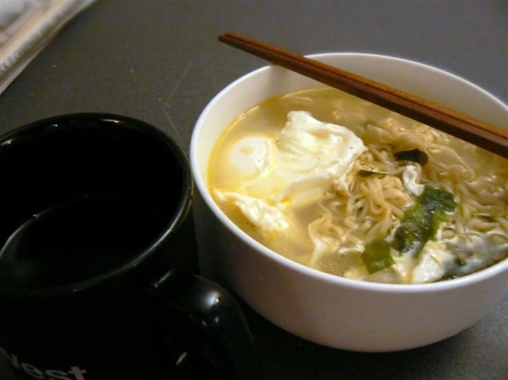 a black coffee cup with chop sticks in it next to a bowl full of chinese noodles and veggies