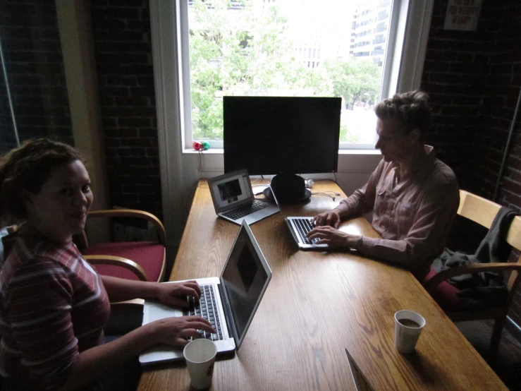 two women sitting at a wooden table with laptops