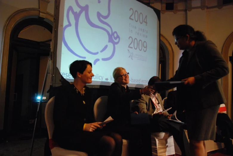 a man stands near a woman in black and other men are seated on chairs