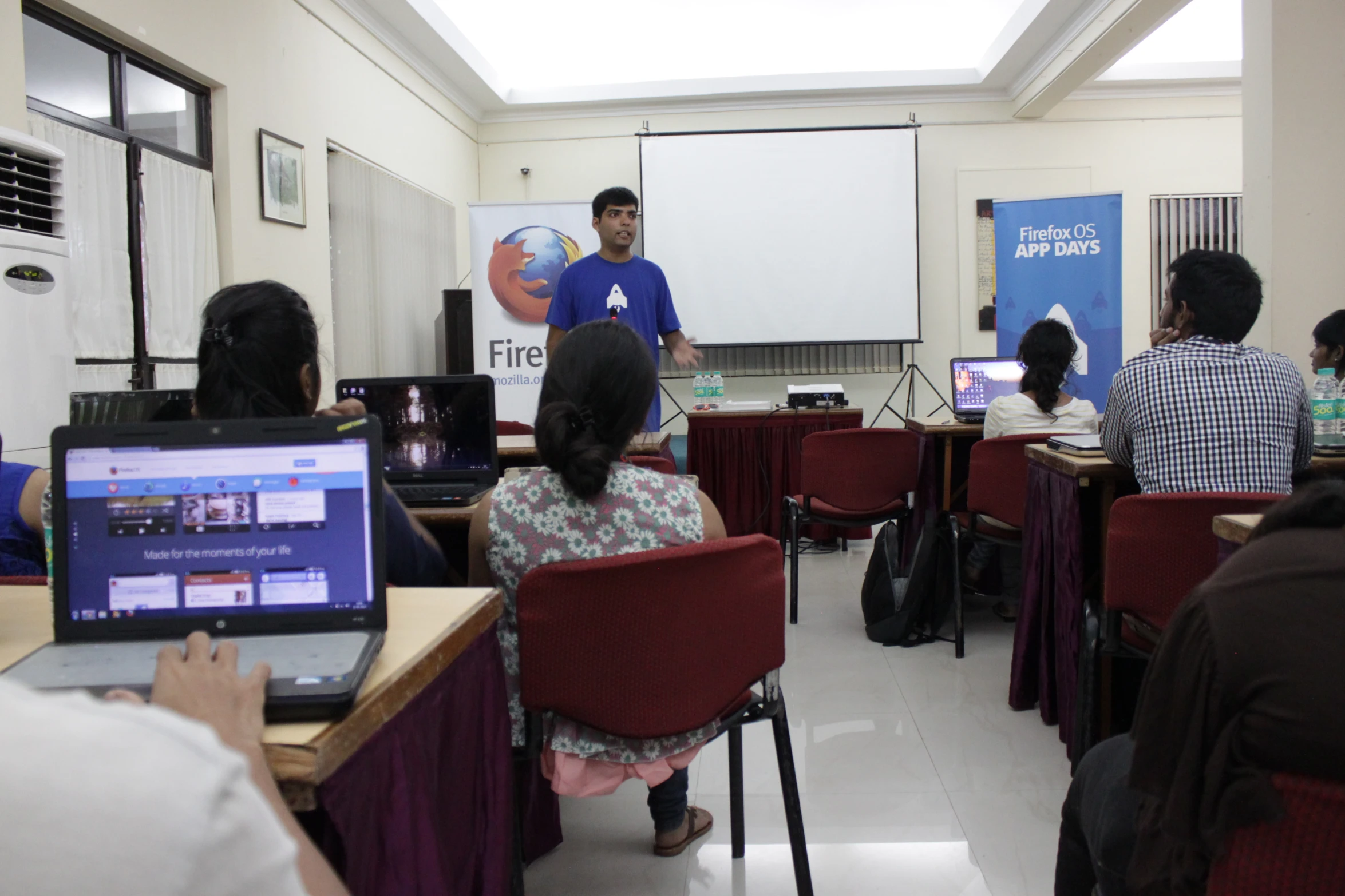 a man standing at the front of a classroom as students sit behind them on laptops