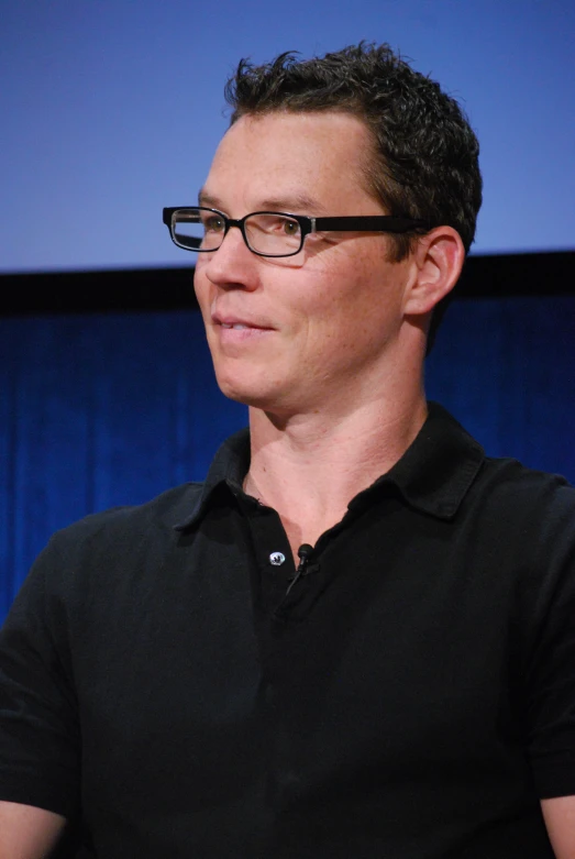 the man is sitting in front of a blue backdrop wearing glasses