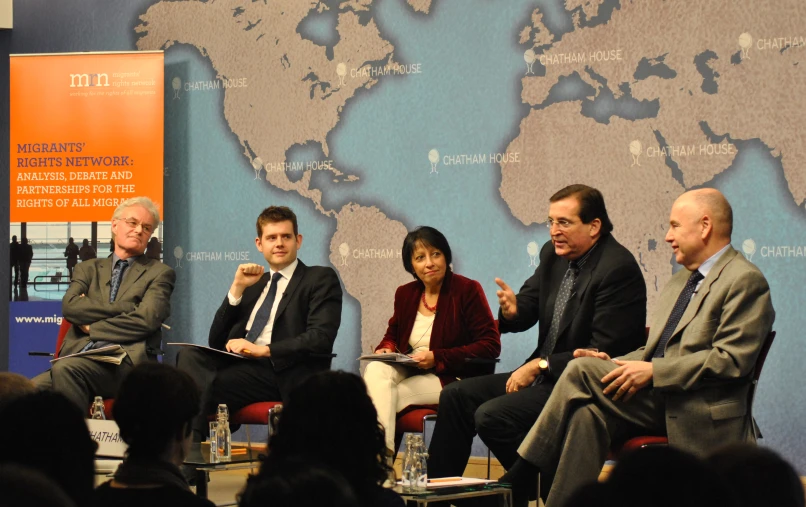 four men and two women sit on chairs talking in front of a world map