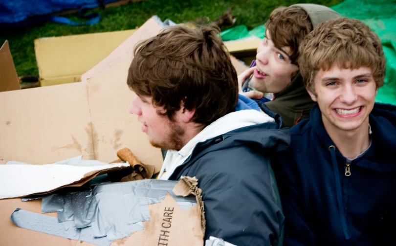 three boys smiling at each other while sitting in cardboard boxes