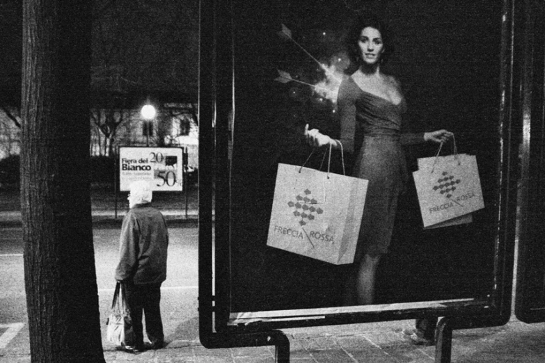 the black and white pograph of a woman on the side of a bus stop with bags