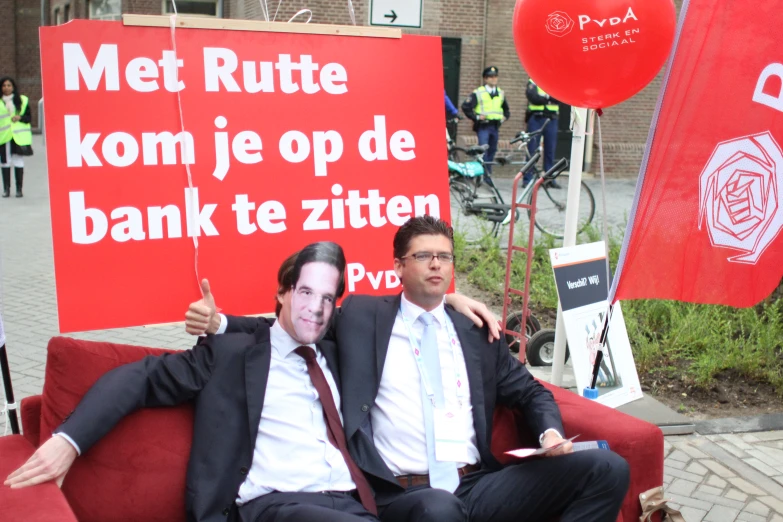 two men sit on a red couch near a sign