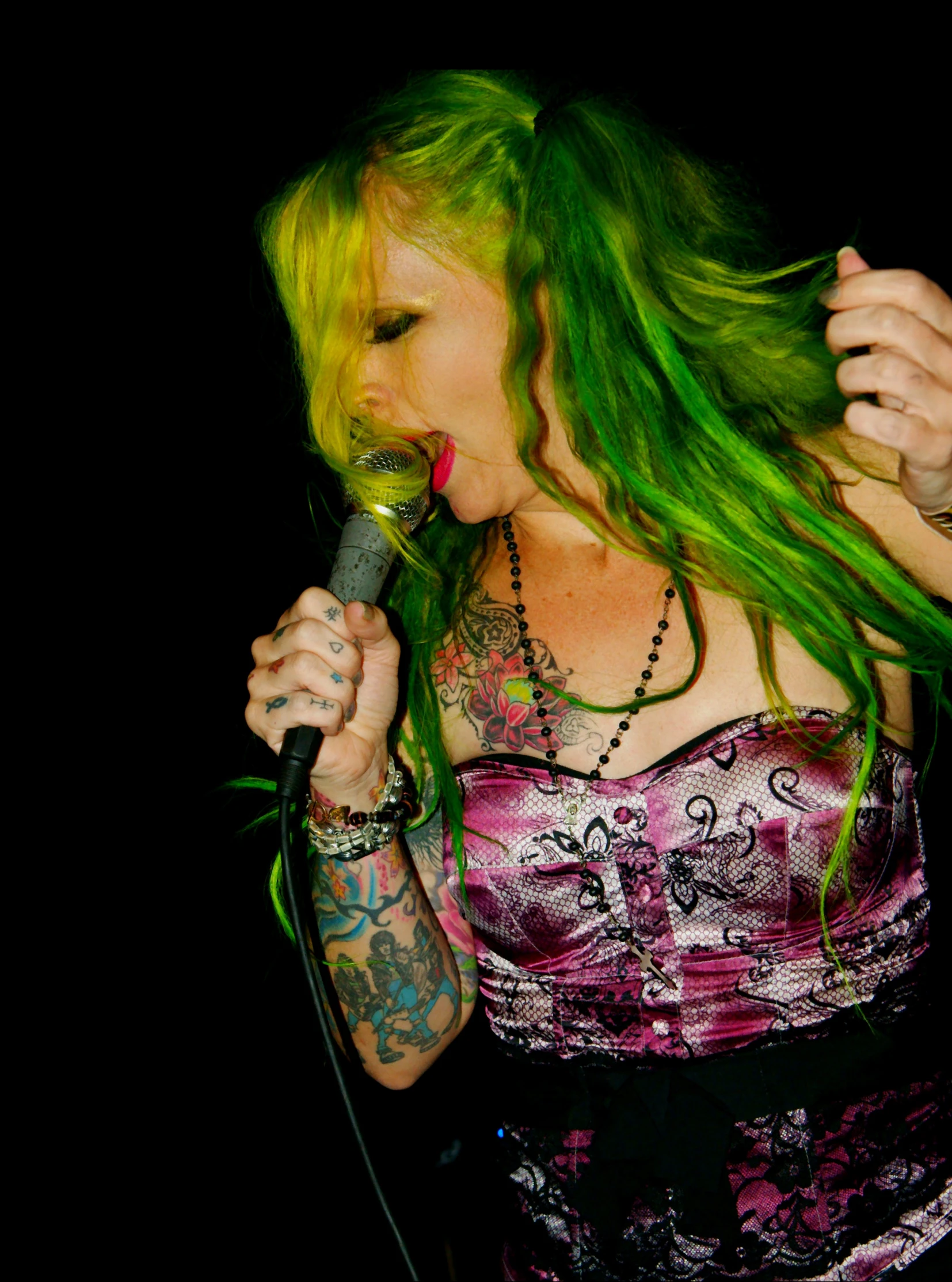 a woman wearing pink and black shirt with long green hair holding a microphone to her mouth