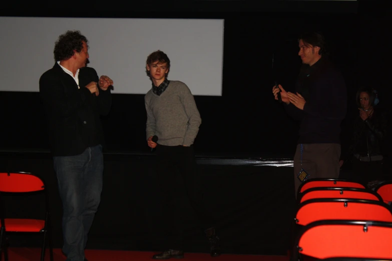 two men in black jackets and two men in dark jackets stand near a screen as people stand in the distance