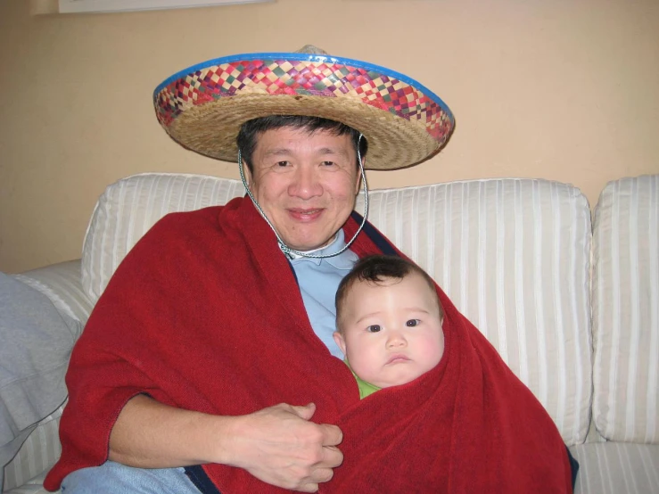 woman sitting on couch covered in red towel holding child with sombrero over head