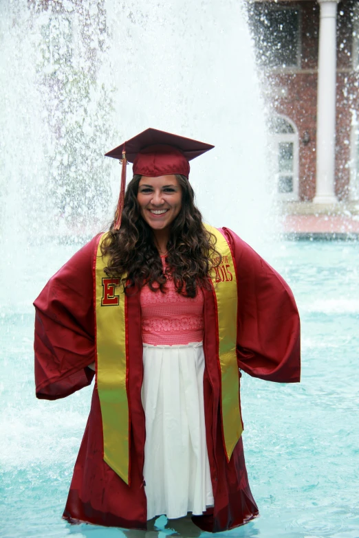 a woman in a red and yellow graduation gown with her hands behind her back, in front of a fountain