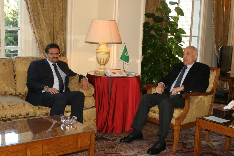 two men in suits sitting on couches next to a lamp