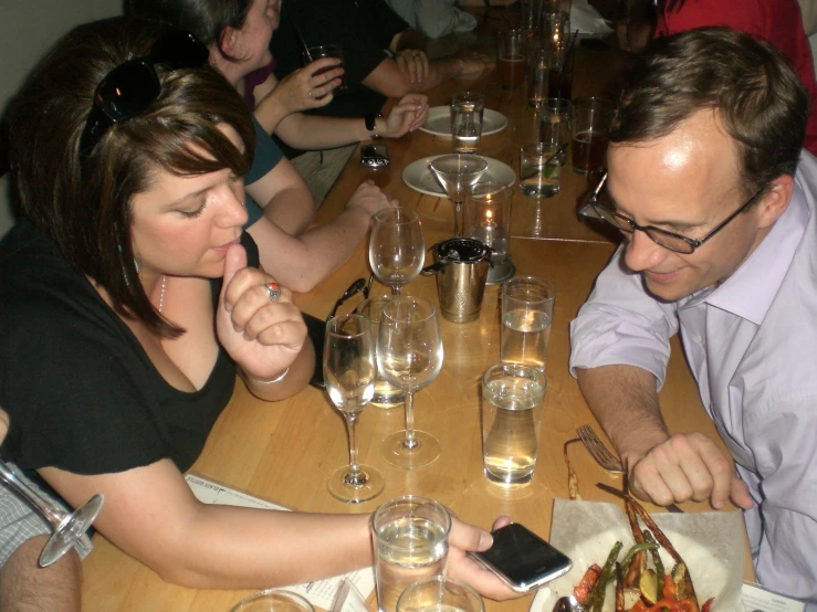 a woman and man sitting at a wooden table using cellphones