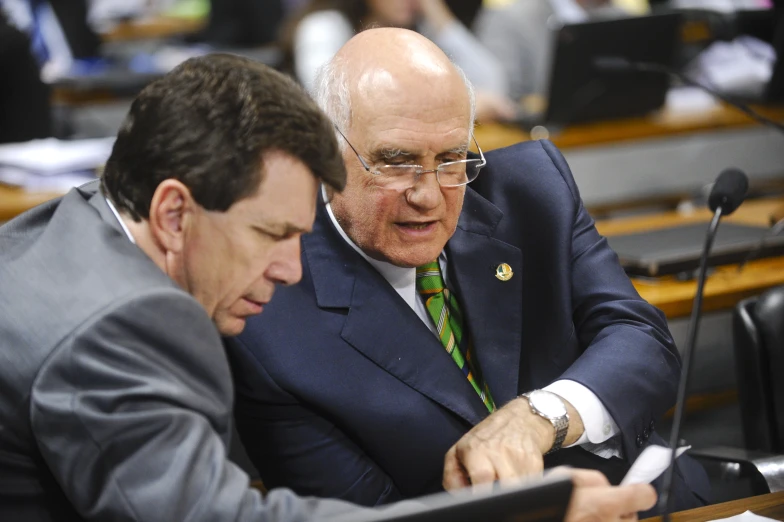 two men in suits sitting at a table and looking at soing