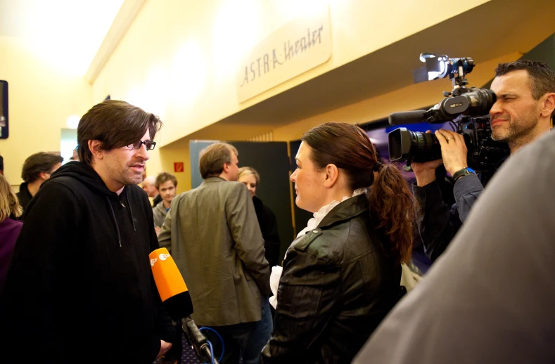 man and woman talking to the media in a room with other people