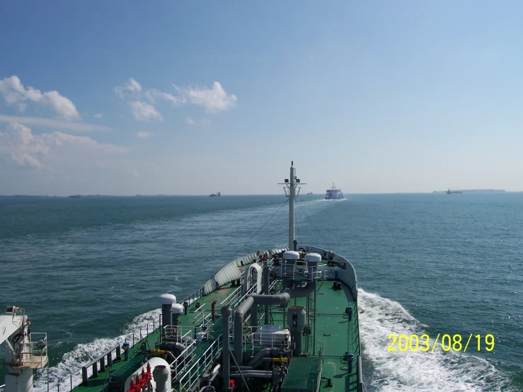 a green boat in the ocean with tugboats and other ships