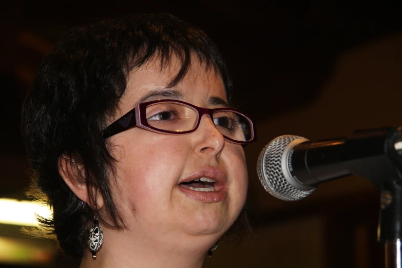 a woman in glasses and a necklace singing into a microphone